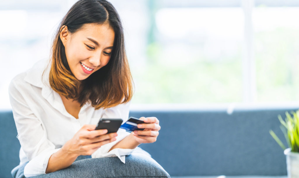 Image depicts a woman sitting on the couch while holding her phone and a credit card