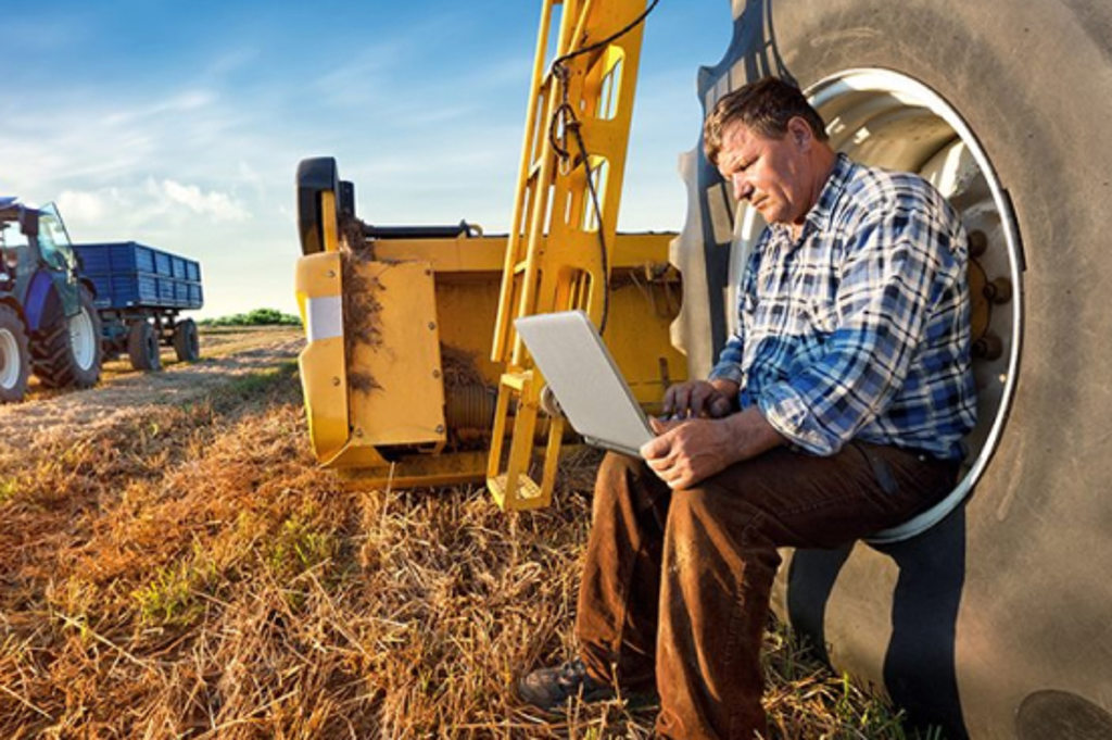 This image depicts farmer sitting on a large combine wheel while he works on a laptop.