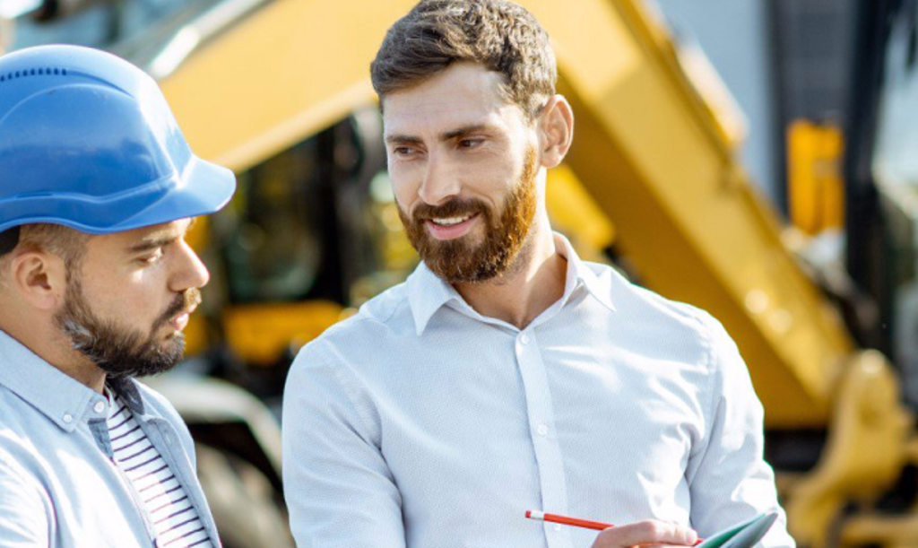 Construction Manager This image depicts a business owner talking to an employee who is wearing a blue hard hat while they stand in front of yellow construction equipmment.