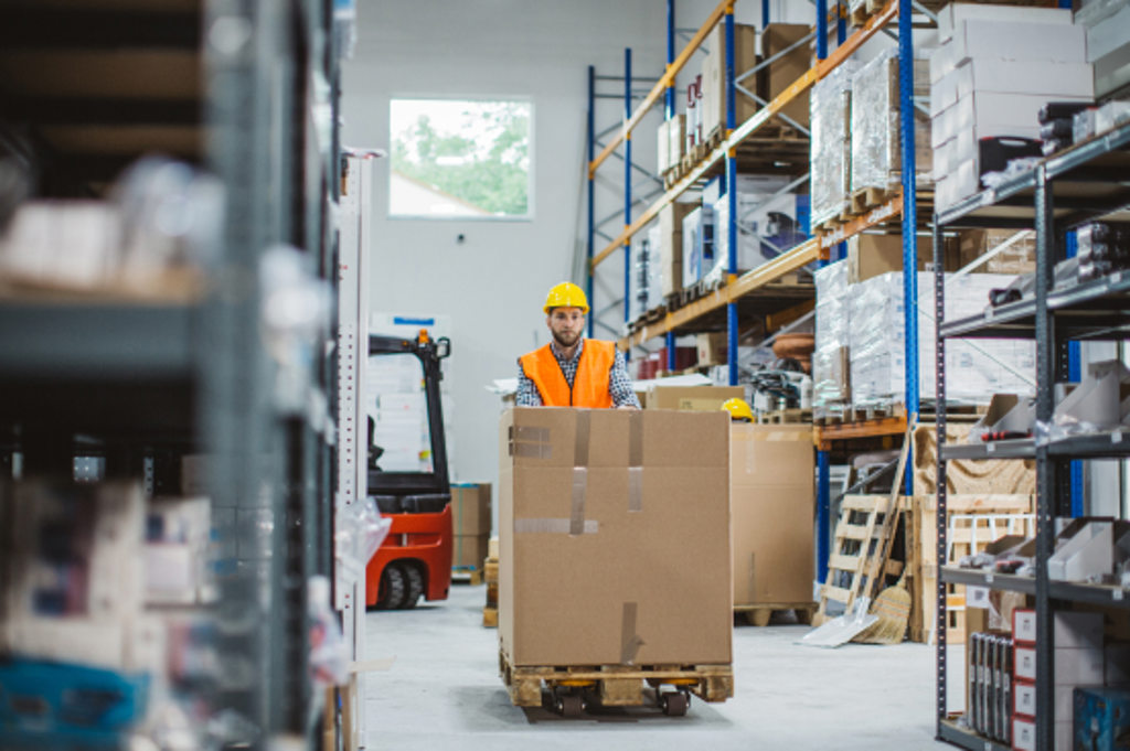 This image depicts an employee in a warehouse wearing a safety helmet and vest while driving a forklift that has a large box on it.