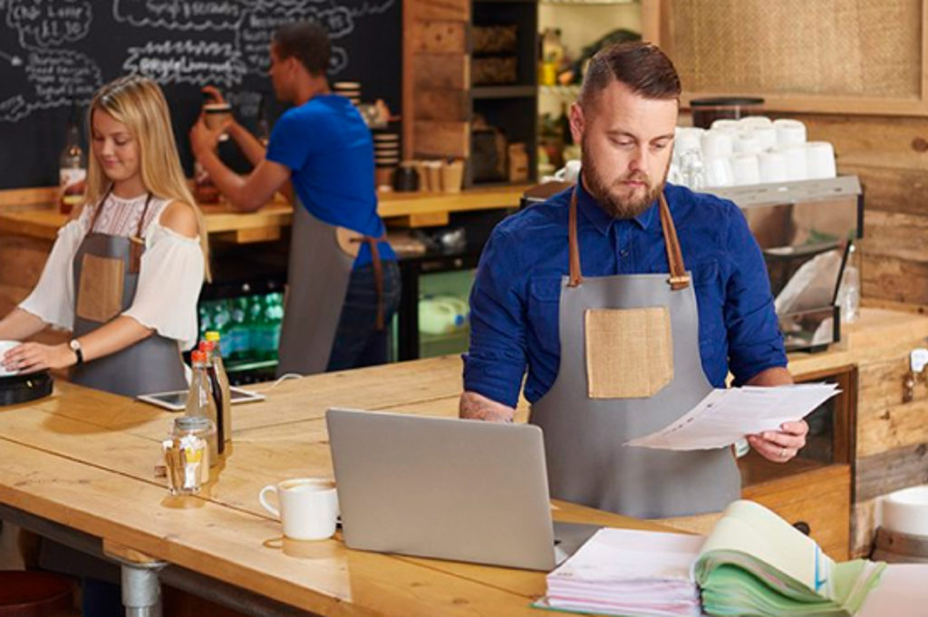 Image depicts the owner of a coffee shop working on a laptop and looking at papers
