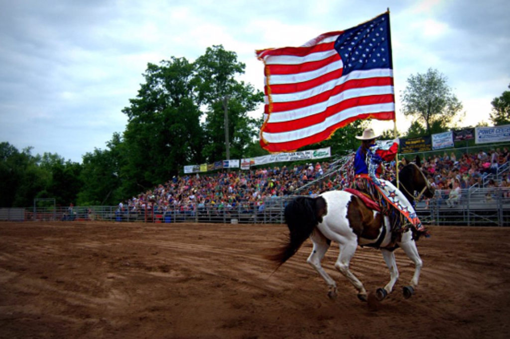 This image depicts a person carrying a large American flag while riding a horse around a ring at a rodeo.