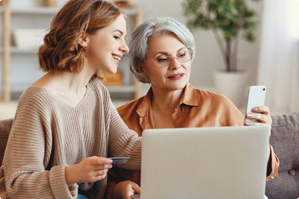 Image depicts a girl and her grandmother sitting on the couch while the granddaughter helps her on her phone and computer